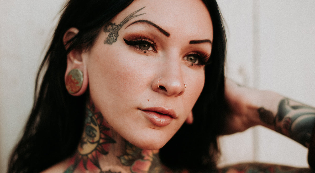 Tattooed woman with a gold-tone nostril hoop, a stud in the other nostril and a green and red stone plug in her lobe