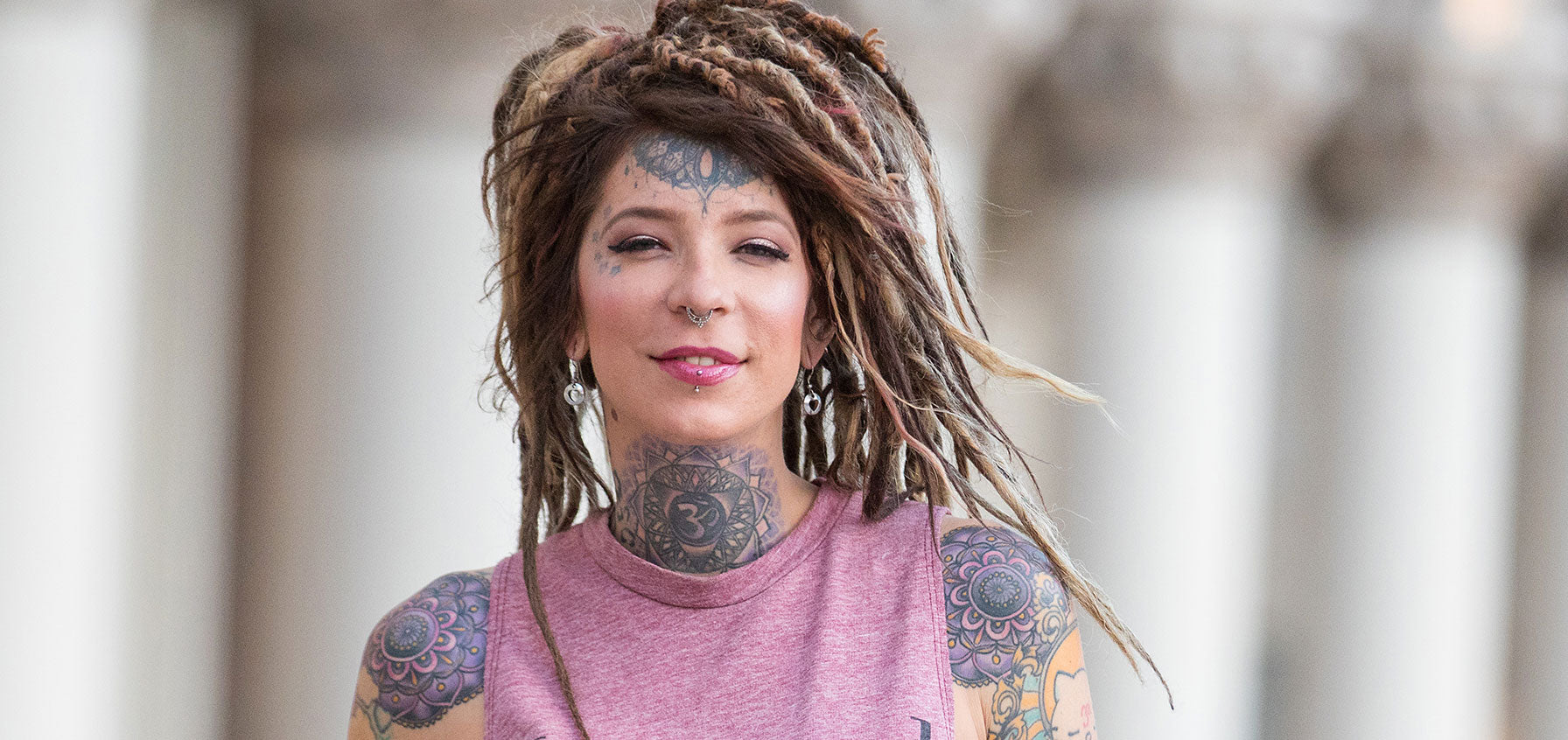 Tattooed woman with long dreadlocks smiling, wearing silver-tone dangle earrings, with tattoos on her forehead and throat
