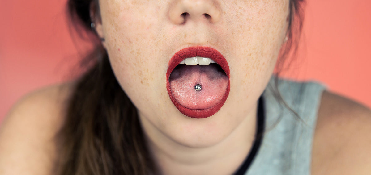 Open mouth with red lipstick showing a center tongue piercing with a silver-tone ball