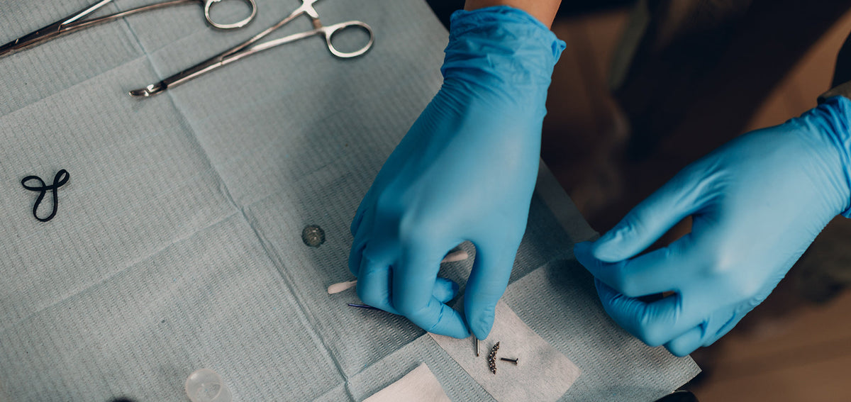 Gloved hands laying out piercing forceps, a labret post and a jeweled curved top on a sterile drape