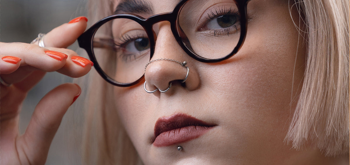 Woman in round glasses with two nostril hoops, a septum ring, a chain across her nose and a labret stud