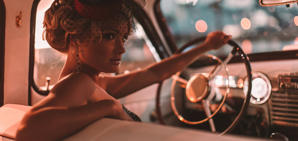 Woman with a curled updo and polka dot veiled hat at the wheel of a vintage car, wearing long beaded drop earrings