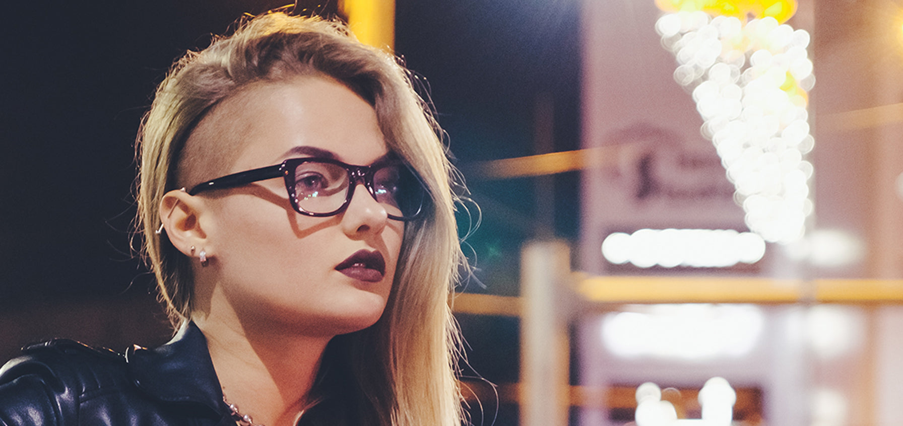 Woman with an undercut and glasses wearing a small stud in her earlobe, with city lights behind her