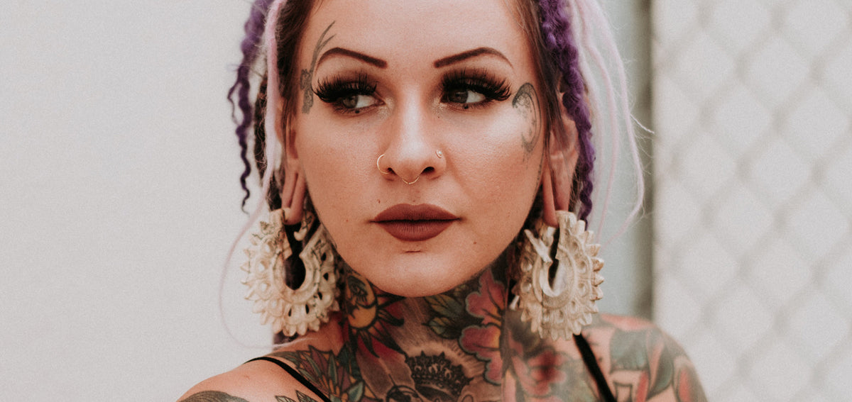 Tattooed woman with purple braids wearing a nostril hoop, a nostril stud, a septum ring and large carved ear weights