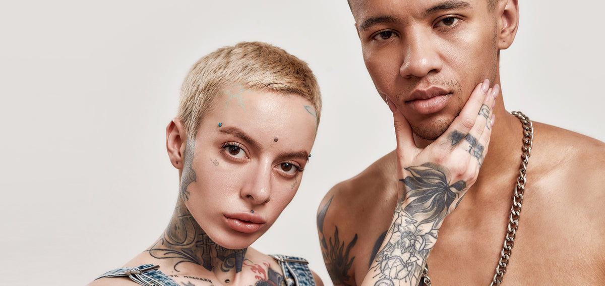 Tattooed person with short blonde hair and studs at the brows, forehead and upper lip, beside a shirtless man in a chain