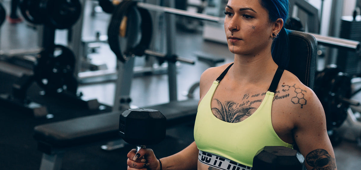 Tattooed woman with blue hair lifting dumbbells in a gym, wearing a nostril ring and hoops in her ear
