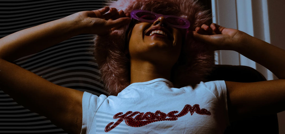 Smiling woman leaning back in a pink faux fur hat and purple sunglasses, wearing a septum ring and a white tee
