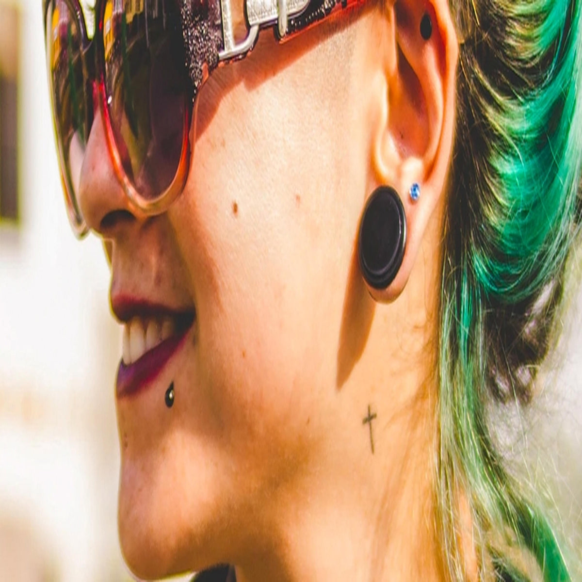 Person in sunglasses with green-streaked hair wearing a black plug, a blue gem stud, an upper ear stud and a labret stud