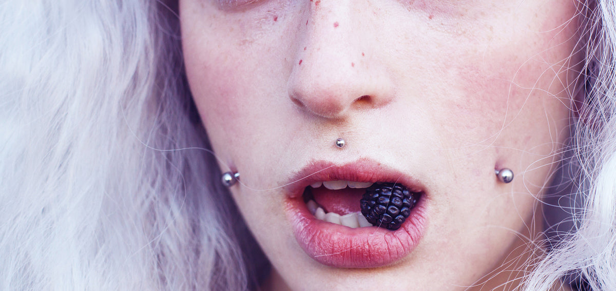 Person with silver hair wearing cheek piercings on both sides and a philtrum stud, holding a blackberry between their lips