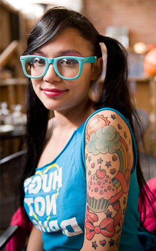 Woman in teal glasses and pigtails with a lip stud and a cupcake and rainbow tattoo sleeve on her upper arm