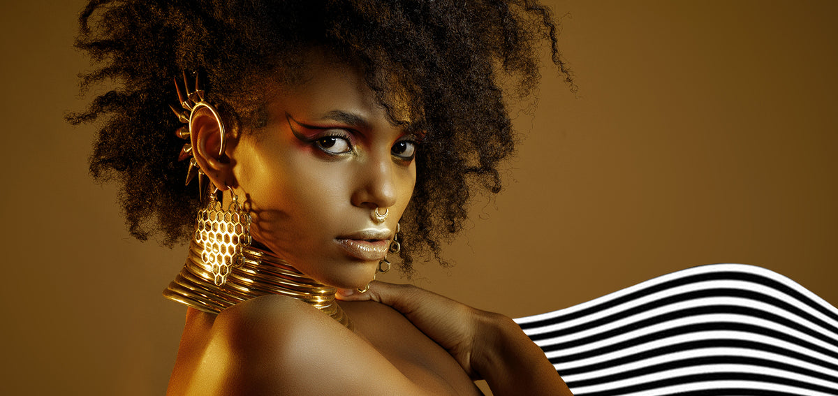 Woman in gold light wearing a spiked gold-tone ear cuff, honeycomb earrings, a septum ring and stacked neck rings
