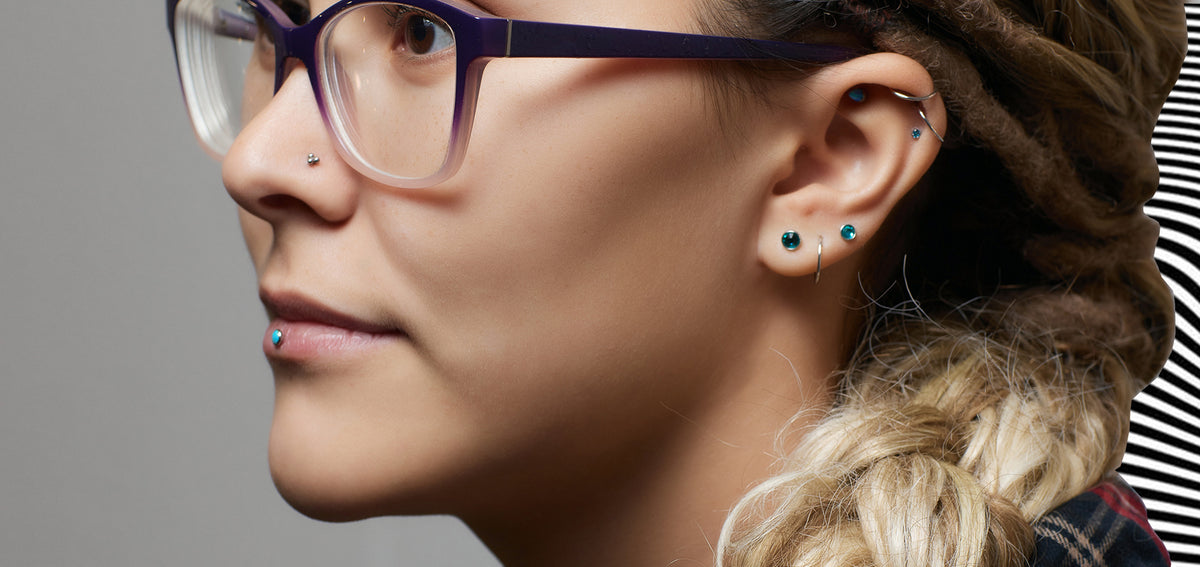 Woman in purple glasses with teal gem studs in her ear and below her lip, thin helix hoops and a nostril stud