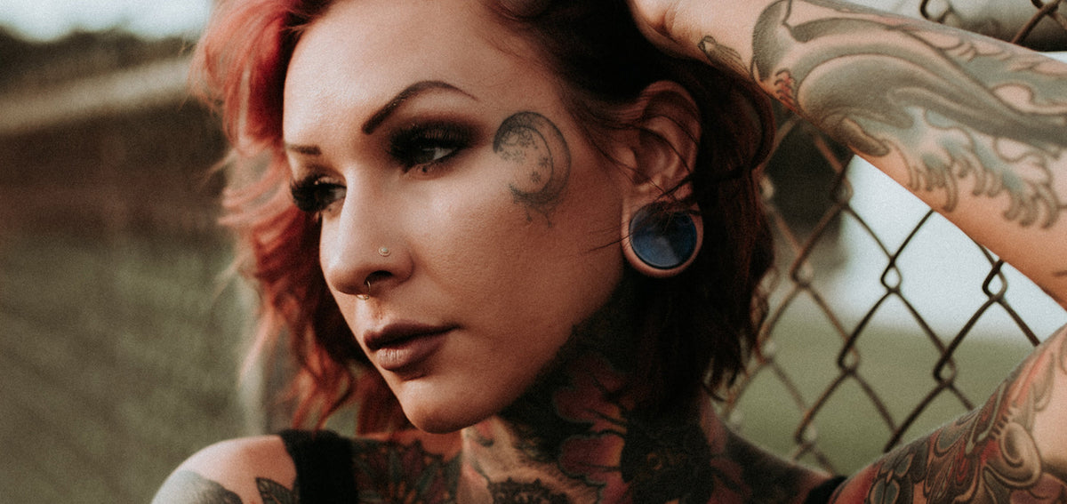 Woman with red hair and a crescent moon face tattoo, wearing a dark blue plug in her stretched lobe and a nostril stud