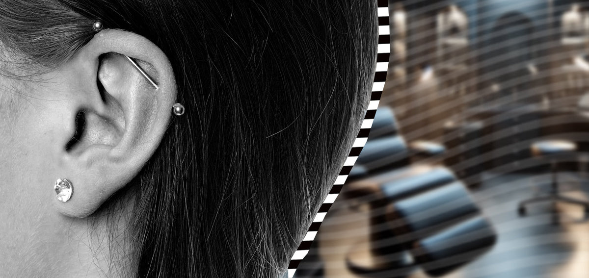Black and white close-up of an ear with an industrial barbell across the upper cartilage and a crystal lobe stud