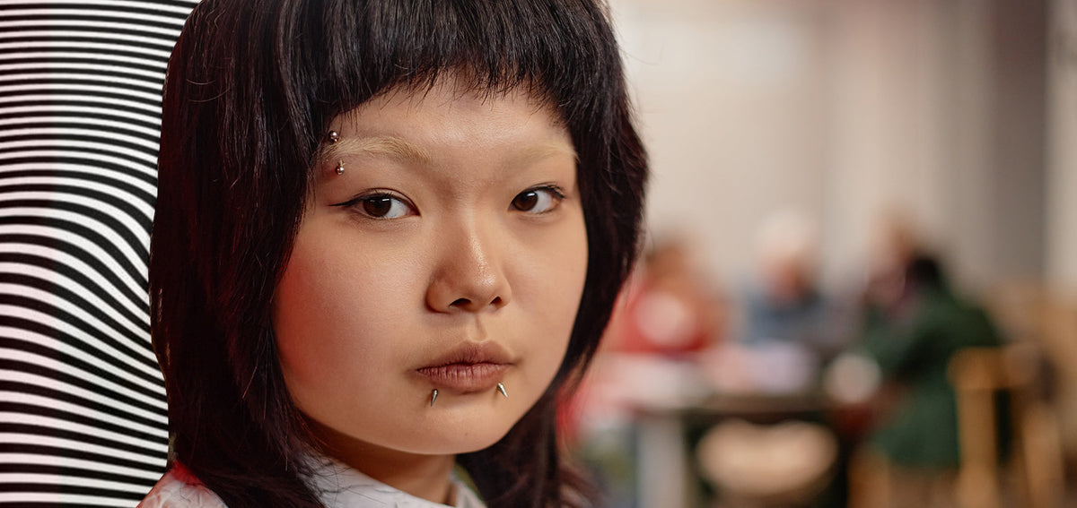 Person with dark bangs wearing a silver-tone eyebrow barbell and studs at both corners of the lower lip