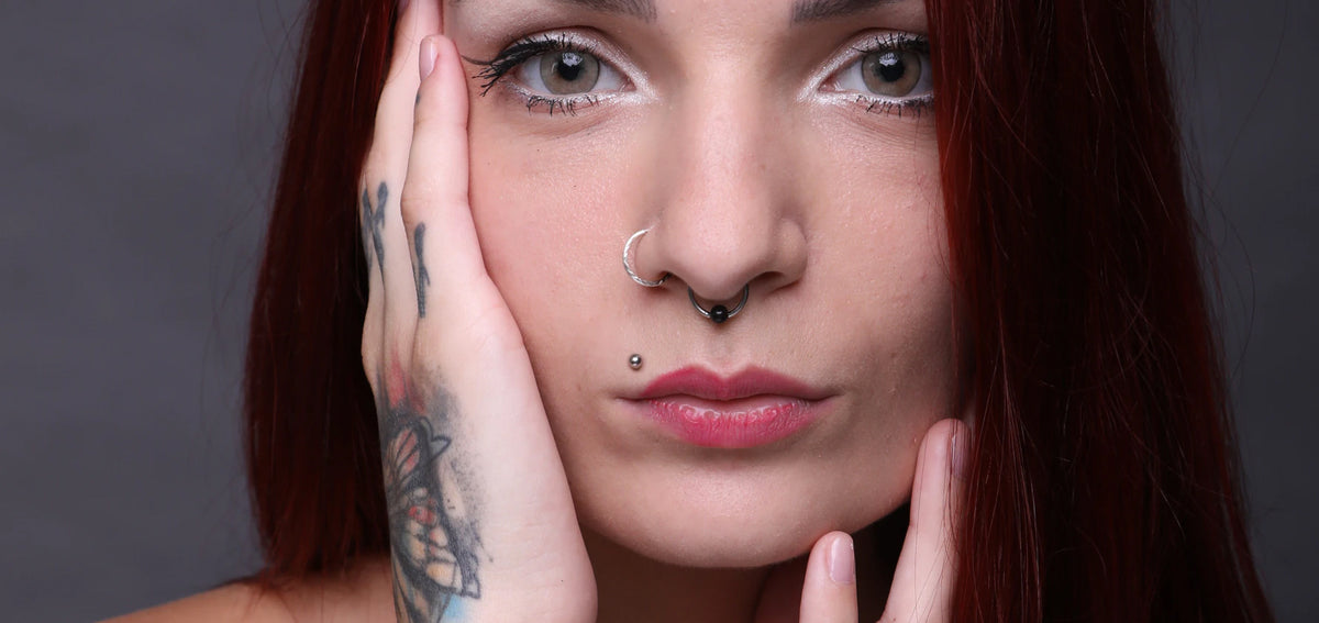 Woman with red hair resting a tattooed hand on her cheek, wearing a nostril hoop, a septum ring and an upper lip stud
