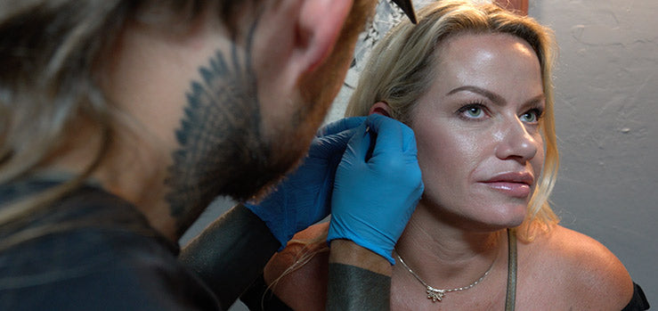 Bearded piercer with a neck tattoo and blue gloves checking a blonde woman's ear before piercing