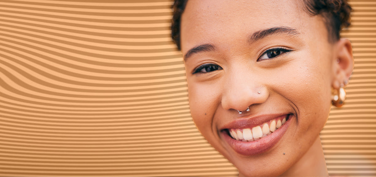 Smiling woman with a silver-tone circular barbell in her septum, a nostril stud and chunky gold-tone hoop earrings