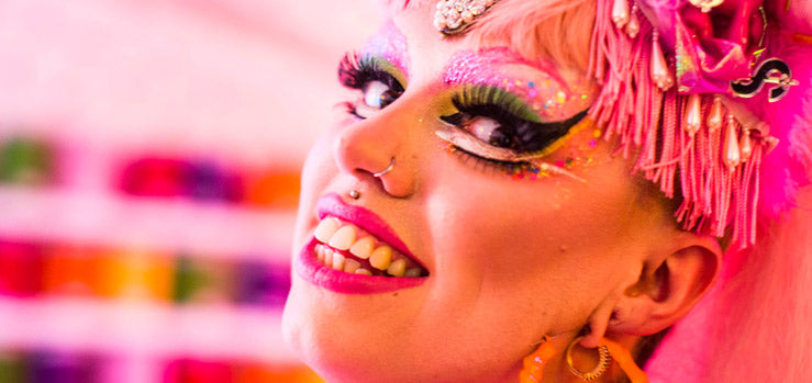 Person in bold glitter makeup and a pink fringed headpiece, wearing a nostril hoop and gold-tone hoop earrings