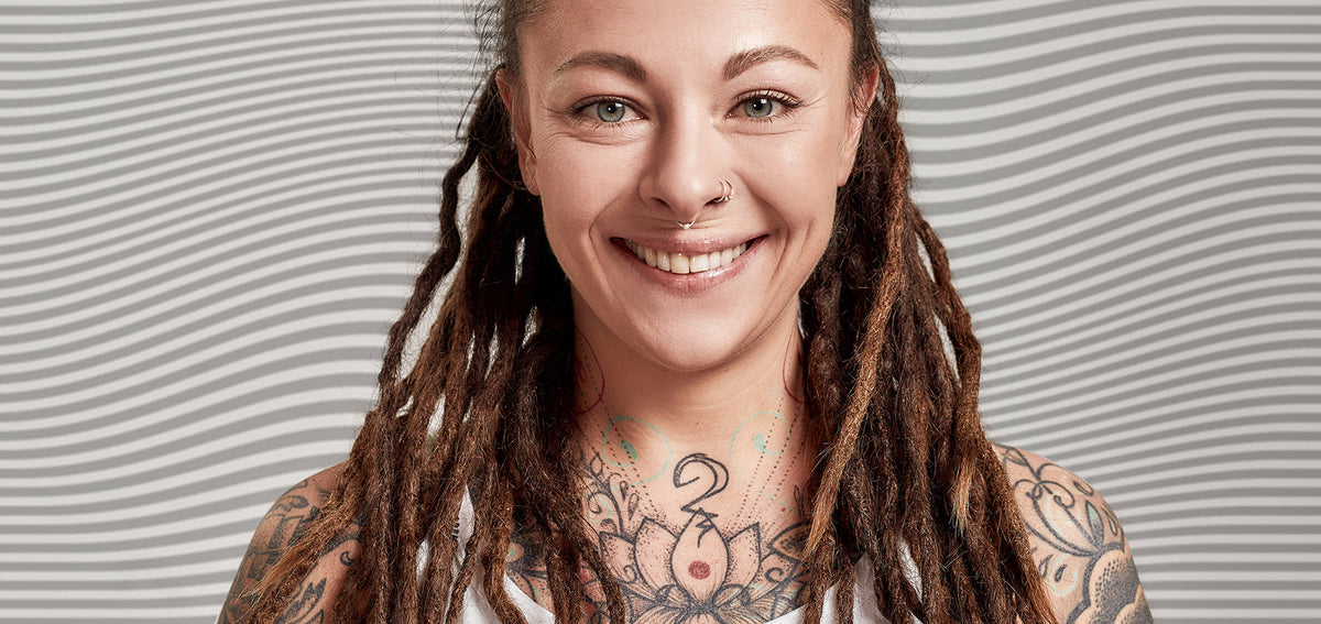 Smiling woman with long dreadlocks and chest tattoos, wearing two small nostril hoops and a septum ring