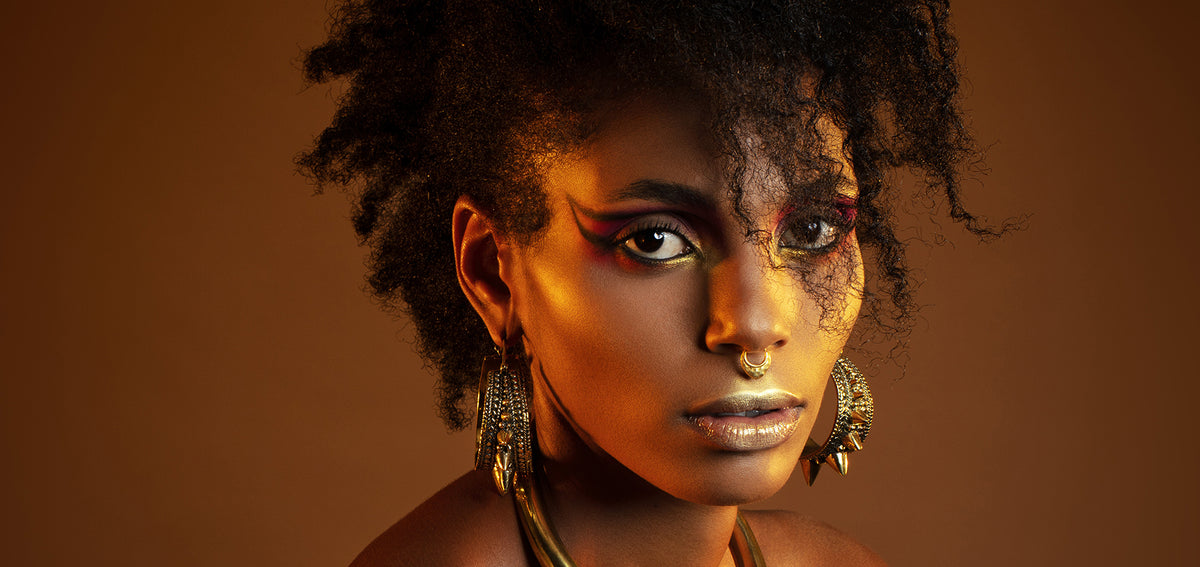 Woman in gold makeup wearing an ornate gold-tone septum ring and large ornate gold-tone earrings