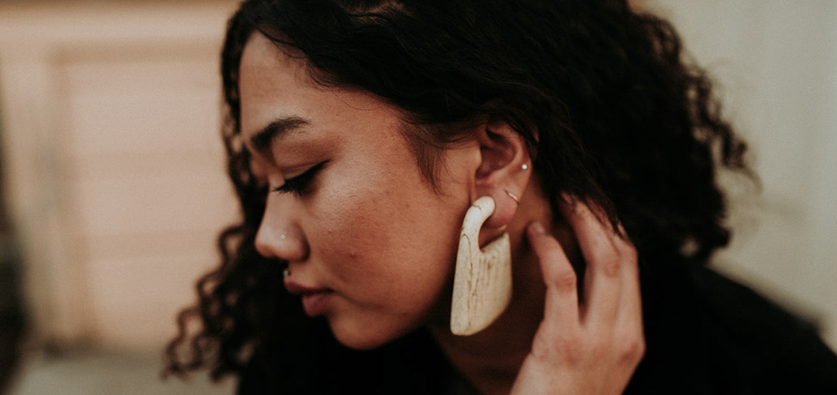 Woman with curly hair wearing a large cream hanger in her stretched lobe, plus a small stud and hoop higher on her ear