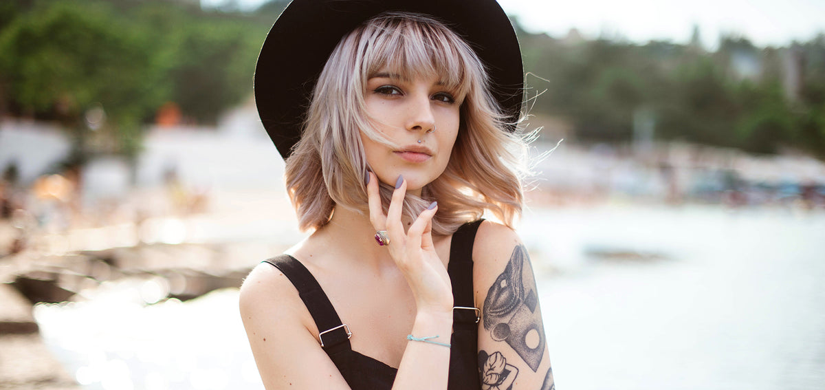 Woman in a black wide-brim hat and black overalls posing by the water, with tattoos on her arm