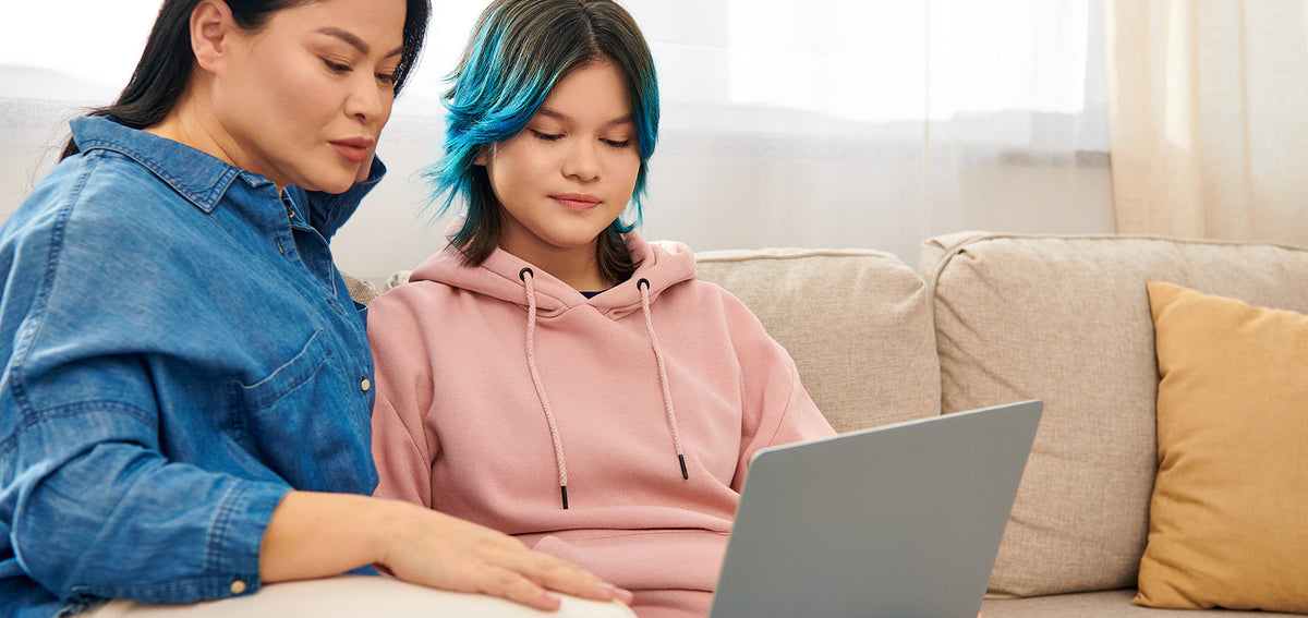 Two people on a couch looking at a laptop together, one with blue-tipped hair in a pink hoodie