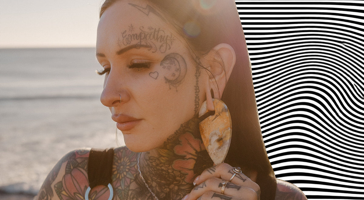 Woman with face tattoos and a nostril ring wearing a large polished stone ear weight in her stretched lobe, at the beach