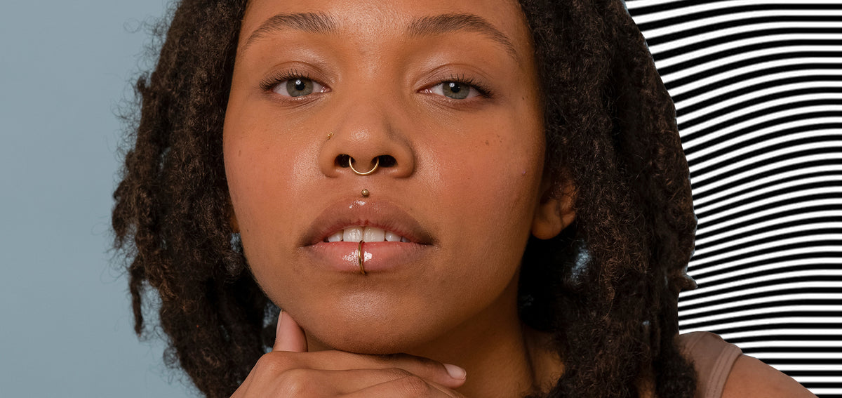 Woman with locs wearing a gold-tone nostril stud, septum ring and medusa stud, plus a thin ring through her lower lip