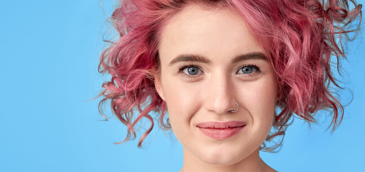 Woman with curly pink hair wearing a thin silver-tone nostril hoop against a blue background