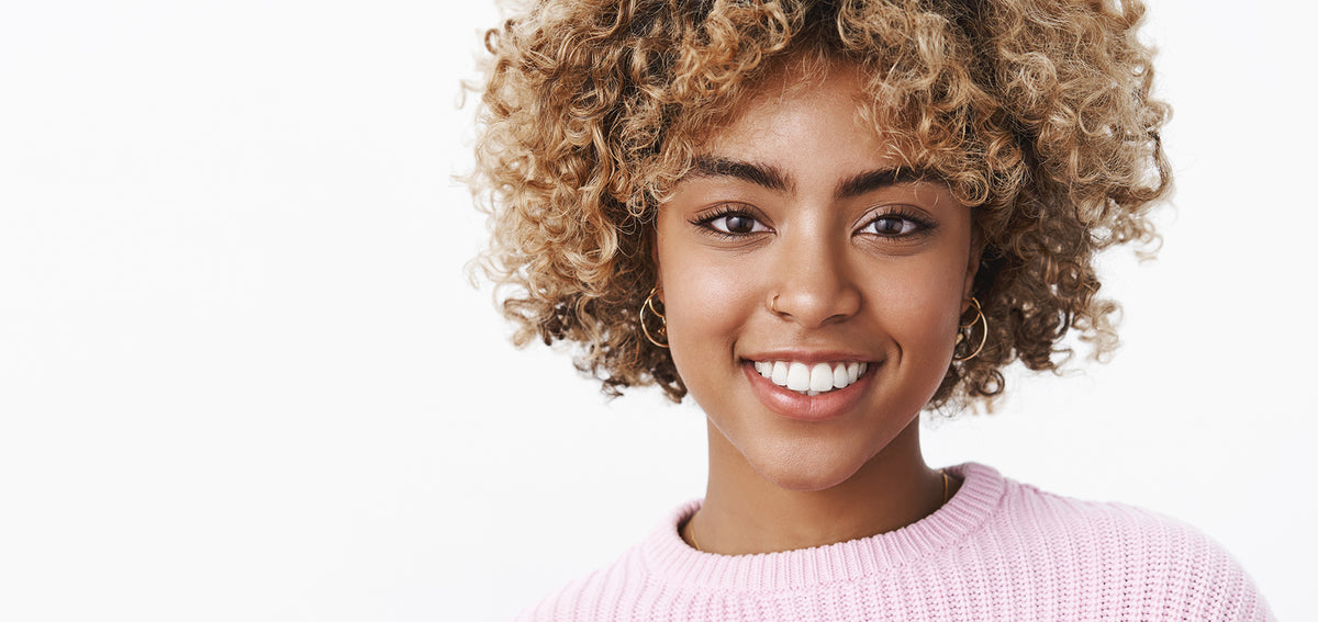 Smiling woman with curly blonde hair, gold-tone hoop earrings and a pink sweater