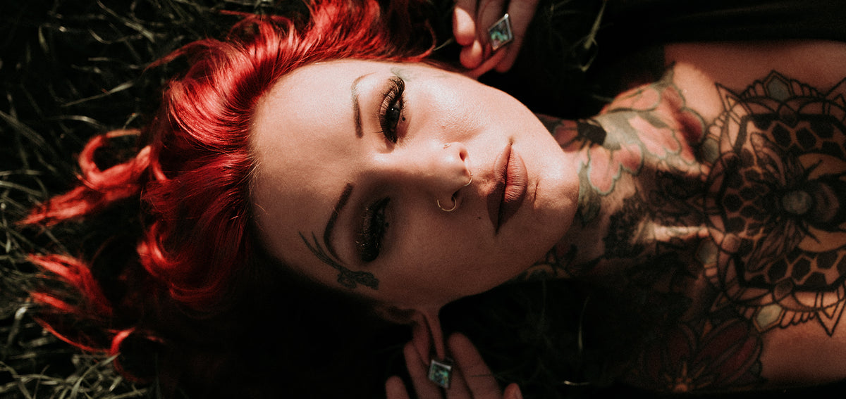 Tattooed woman with red hair lying in the grass, wearing two gold-tone hoops in one nostril