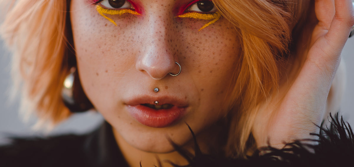 Freckled woman with orange hair and yellow eyeliner, wearing a vertical philtrum barbell and a nostril hoop