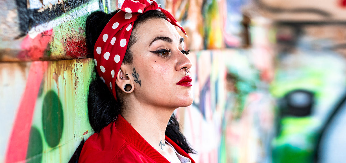 Woman in a red polka dot head scarf by a graffiti wall, wearing a black plug in her lobe and a septum ring