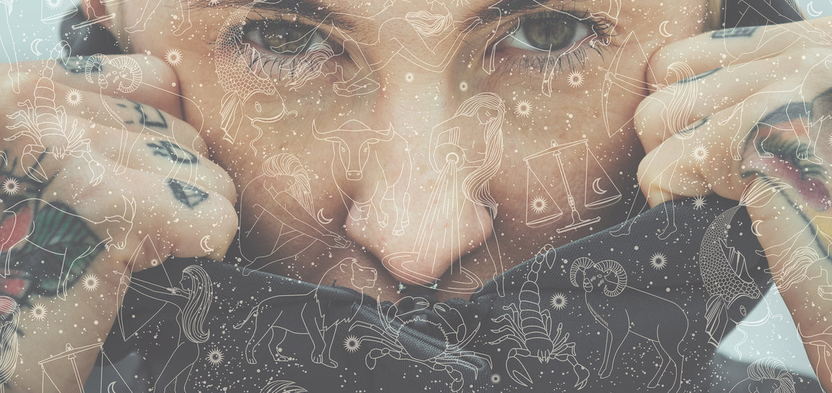 Person with tattooed hands peering over a dark cloth, overlaid with line drawings of zodiac signs