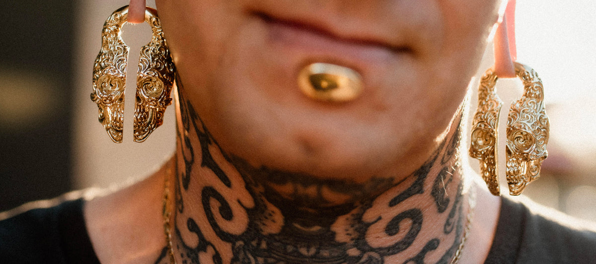 Ornate gold ear weights hanging from stretched earlobes, worn with a gold lip labret